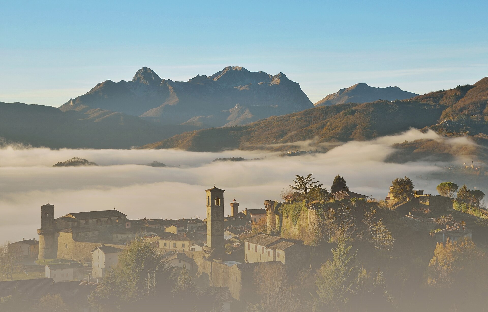 The walled Rocca of Castiglione di Garfagnana at dawn above valley fog, with the Apuan Alps behind