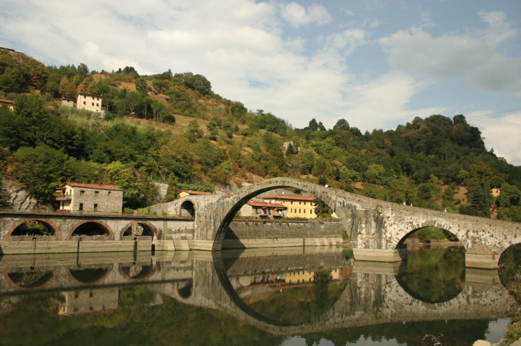 Ponte della Maddalena's tall stone arch reflected in the still Serchio river at Borgo a Mozzano, wooded hills behind