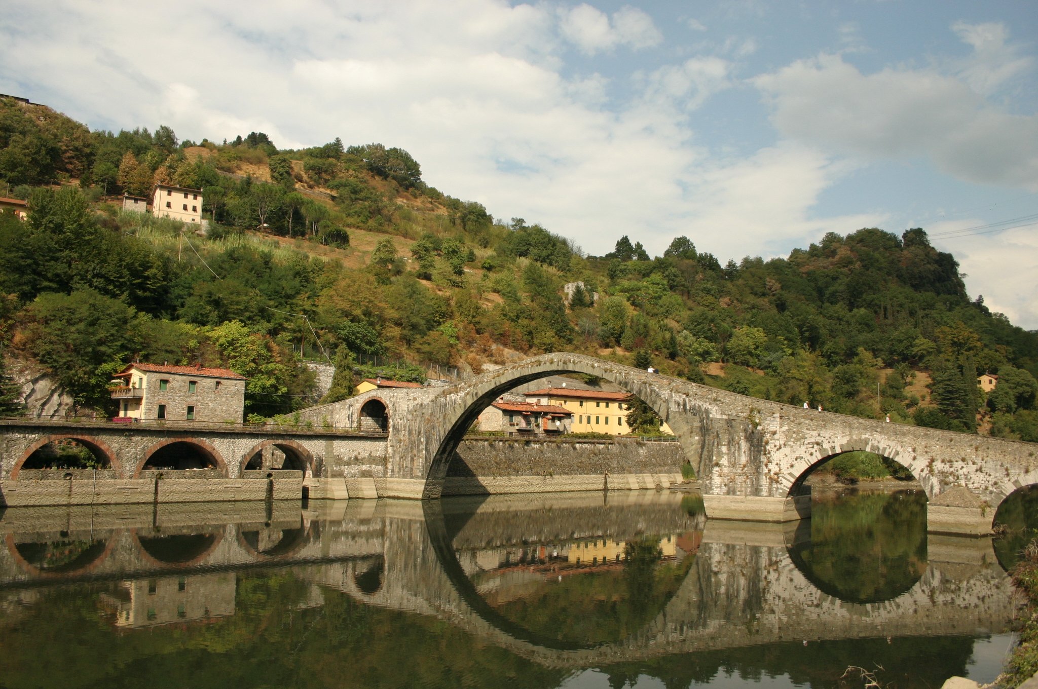 Ponte della Maddalena's tall stone arch reflected in the still Serchio river at Borgo a Mozzano, wooded hills behind